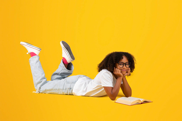 Positive smart teen black curly girl in white t-shirt, glasses reads book, lies on floor, isolated on yellow studio background. Study, knowledge, hobby, rest and relax, ad and offer - Foto, Bild