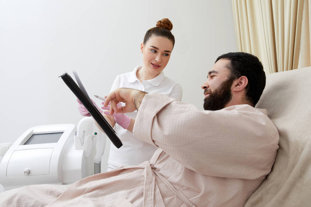 Senhora sorridente mostra papel para homem cliente sentado em poltrona confortável na clínica cosmetológica - Foto, Imagem