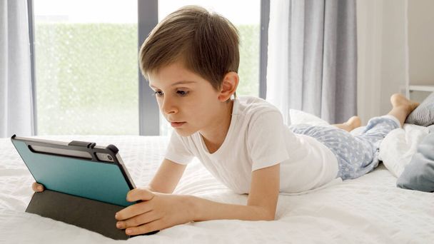 Νεαρό αγόρι απολαμβάνει χρόνο οθόνης στο κρεβάτι του, χρησιμοποιώντας έναν υπολογιστή tablet για να εξερευνήσετε τον ψηφιακό κόσμο. Παιδί που χρησιμοποιεί gadget, εκπαίδευση και ανάπτυξη - Φωτογραφία, εικόνα