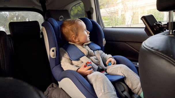 Little baby by sitting in the safety child car seat and looking out of the open window. Concept of kids traveling, baby safety, children going by car - Photo, Image