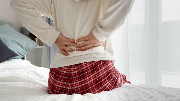 Een jonge vrouw in een pyjama die op het bed zit en haar rugpijn masseert. Een concept van gezondheidsproblemen, pijnbestrijding en de behoefte aan rust. - Foto, afbeelding