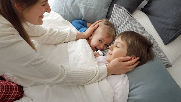 Mladá starostlivá matka přikrývající své dva syny ležící v posteli přikrývkou a hladící je. Koncept rodinného štěstí, relaxace doma, zábava v posteli, rodiče a veselé děti. - Fotografie, Obrázek