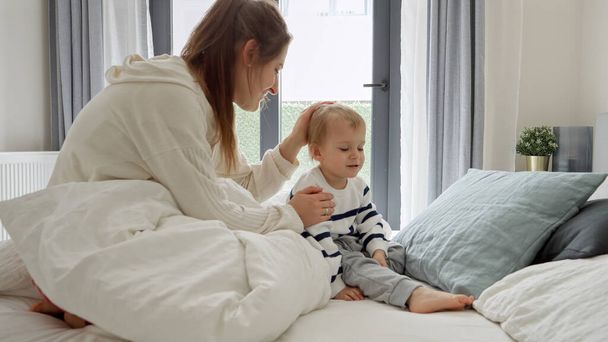 Young mother caressing and soothing her upset toddler son lying in bed - Photo, Image