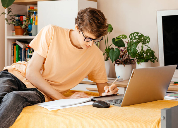 Сконцентрированный молодой подросток, работающий над ноутбуком на диване дома. Улыбающийся мальчик делает домашнее задание с помощью компьютера. Дистанционное, онлайн образование для детей - Фото, изображение