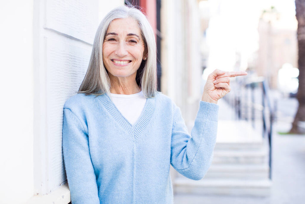 senior retired pretty white hair woman smiling cheerfully, feeling happy and pointing to the side and upwards, showing object in copy space - Foto, Imagem