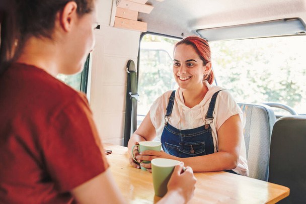 Two young women drinking coffee and having a great time in a handmade camper van. Van road trip holiday and outdoor summer adventure. Nomad lifestyle concept - Photo, Image