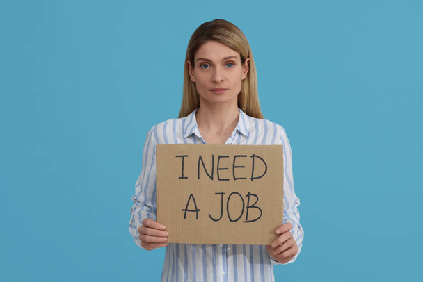 Unemployed woman holding sign with phrase I Need A job on light blue background - Valokuva, kuva
