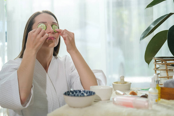 Young woman in bathrobe putting fresh cucumber on her eye while doing beauty treatment at home.	 - Foto, immagini