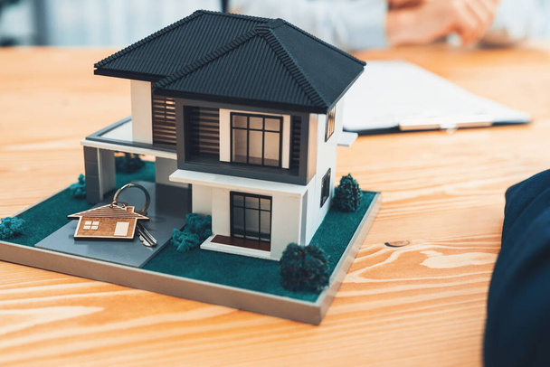 Model domu je zobrazen na dřevěném jednacím stole s rozmazaným pozadím realitního makléře a klient diskutovat o podmínkách úvěru na bydlení nebo nájemní smlouvy. Entita - Fotografie, Obrázek