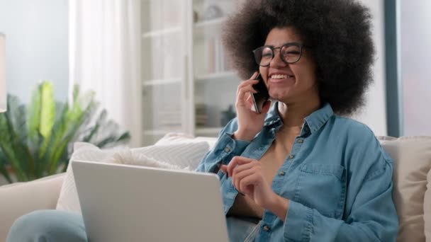 Mulher afro-americana falando telefone celular trabalhando com laptop fazer pedido on-line reserva entrega de alimentos no sofá em casa senhora falando chamada de negócios discutir projeto de computador conversa amigável - Filmagem, Vídeo