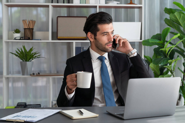 勤勉なビジネスマンは、携帯電話で現代の勤勉なオフィスワーカーのライフスタイルの概念として彼のオフィスでラップトップで作業しながら、クライアントと電話で話を忙しい。フェルベント - 写真・画像