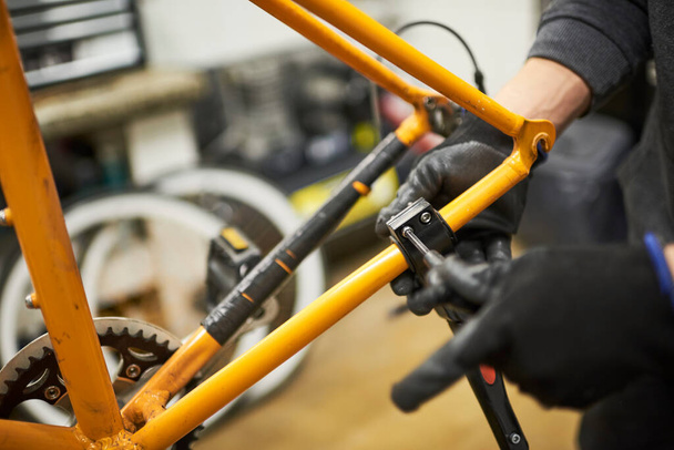 Επισκευή ποδηλάτου: χέρια αγνώριστου προσώπου με γάντια που αποσυναρμολογούν πορτοκαλί ποδήλατο στο εργαστήριό του. Επιλεκτική σύνθεση εστίασης. - Φωτογραφία, εικόνα