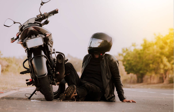 Мотоцикліст сидить поруч зі своїм мотоциклом. Мотоцикліст сидить і спирається на свій мотоцикл на асфальті
 - Фото, зображення