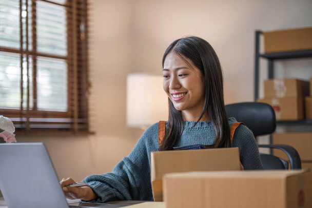 Boldog fiatal ázsiai nő startup kisvállalkozás szabadúszó holding csomag doboz és számítógép laptop és ül a széken, Online marketing csomagolás szállítási koncepció.. - Fotó, kép