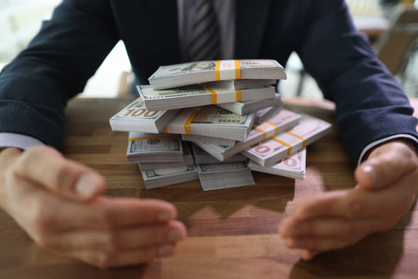 Greedy man in formal suit closes dollars pile with hands at desk in office. Businessman protects large money amount. Financial security - Foto, Bild