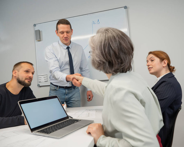 The boss makes a presentation to subordinates at the white board. Caucasian man shaking hands with middle aged woman - Fotoğraf, Görsel