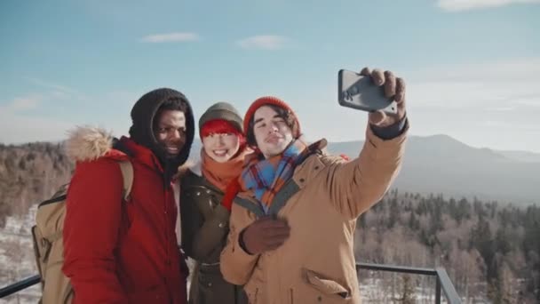Fiatal férfi gazdaság smartphone csinál szelfi a etnikailag változatos barátok, miközben áll a megfigyelő platform a hegy tetején télen nap - Felvétel, videó