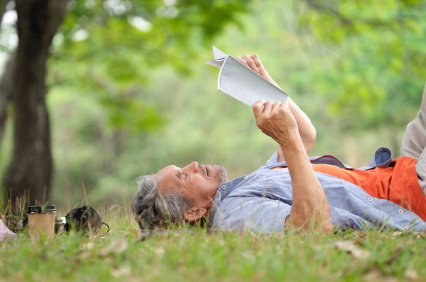 old mature male relax outside in nature garden,happy senior man smile,casual lying on his back on grass reading a book under a tree outdoor in the summer park,concept elderly people lifestyle,resting - Photo, image