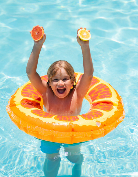 Little kid boy floating in a swimming pool on summer vacation. Kid with orange in the pool. Summer fruits. Happy kid playing with swim ring in swimming pool - Фото, изображение