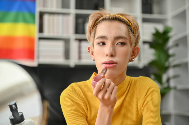 Un jeune blogueur de beauté gay asiatique attrayant appliquant du rouge à lèvres sur sa lèvre, se préparant à la maison. Concept de style de vie LGBTQ + - Photo, image