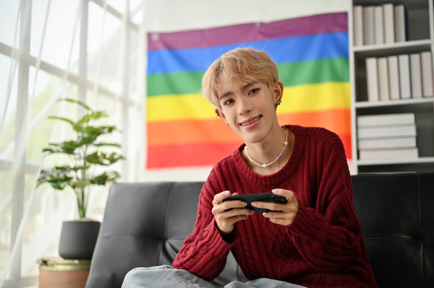 Houkutteleva ja hymyilevä nuori aasialainen homo mies istuu sohvalla älypuhelin käsissään, rentoutuen olohuoneessaan.. - Valokuva, kuva