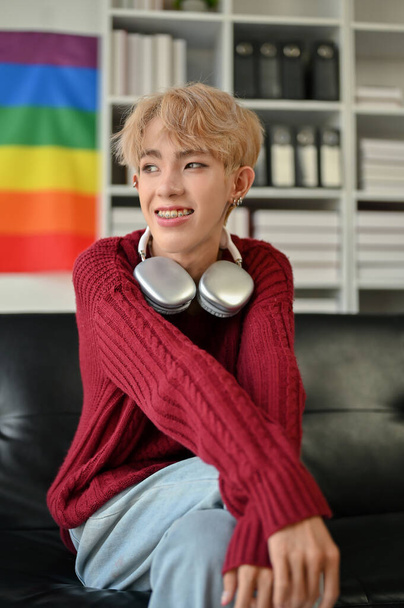 A portrait of an attractive and happy young Asian gay man in casual clothes sits on a sofa in his modern living room. LGBTQ+ lifestyle concept - Photo, Image