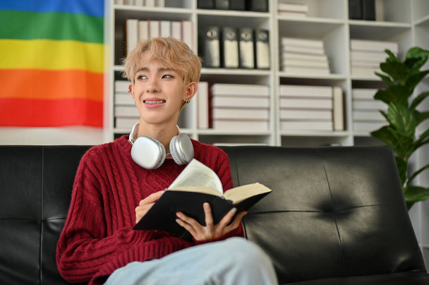 Un giovane gay asiatico felice sta guardando fuori dalla finestra e sognando ad occhi aperti mentre legge un libro sul divano nel suo soggiorno. Stile di vita concetto - Foto, immagini