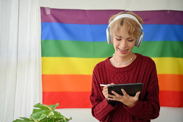 Szczęśliwy młody azjatycki gej w luźnych ciuchach słucha muzyki przez słuchawki i używa tabletu stojąc w pokoju z flagą LGBT za sobą na ścianie. - Zdjęcie, obraz