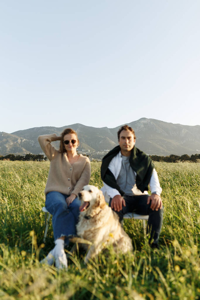 夏の日没の日に羽の草畑の椅子に座って、犬と若い幸せなカップルは、カメラを楽しみにして、深刻な. - 写真・画像