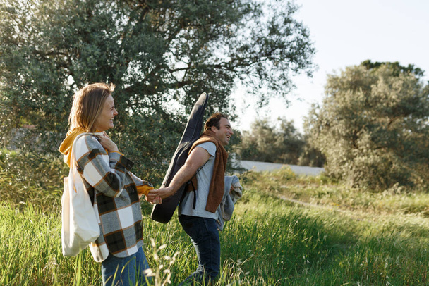 Young middle age couple walking along happily talking in park. Mature couple in summer park. Man and woman in casual clothes with guitar and picnic bag outside in spring Cyprus nature. - Photo, Image