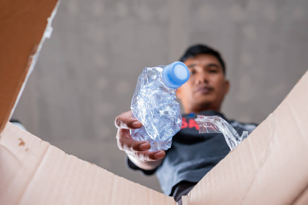 Een man verzamelt en stopt recycleerbare plastic flessen in een kartonnen doos - Foto, afbeelding
