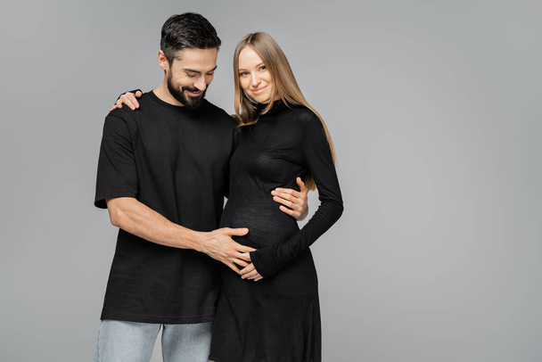 Positive bearded man in black t-shirt touching belly of fair haired pregnant wife in stylish dress and hugging each other while standing isolated on grey, growing new life concept - 写真・画像
