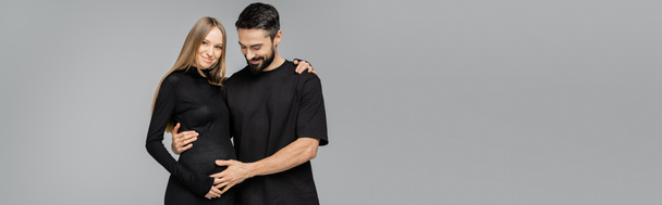 Cheerful bearded man in black t-shirt touching belly of fair haired and pregnant stylish wife in dress and hugging isolated on grey, growing new life concept, copy space, banner  - Photo, Image