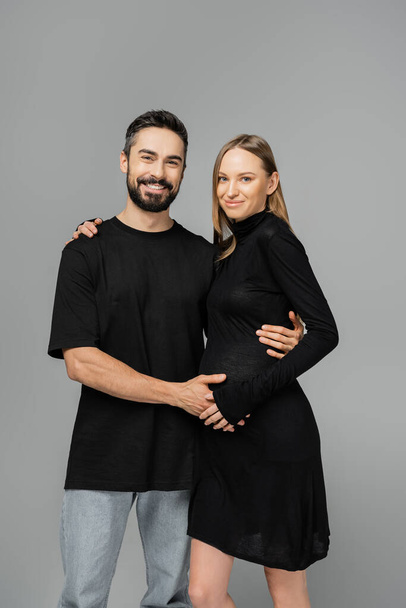 Positiv schwangere Frau in stylischem schwarzen Kleid blickt in die Kamera und umarmt bärtigen Ehemann in T-Shirt und Jeans, während sie isoliert auf grauem, wachsendem neuen Lebenskonzept steht - Foto, Bild