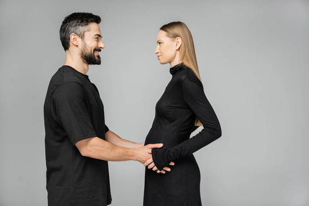 Side view of positive bearded man in t-shirt touching belly of pregnant wife in stylish black dress and looking at each other while standing isolated on grey, growing new life concept - 写真・画像