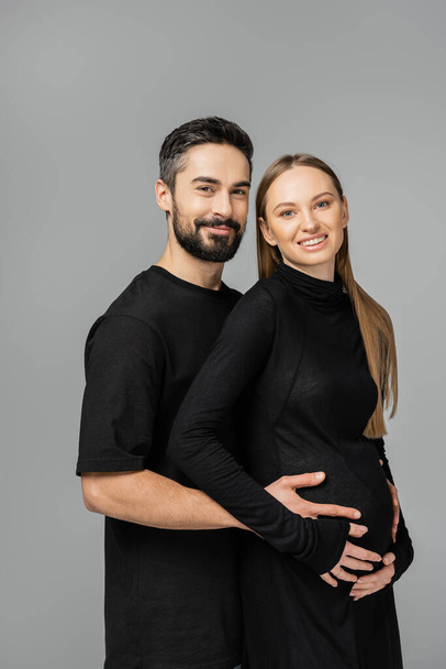 Porträt eines bärtigen Mannes im schwarzen T-Shirt, der in die Kamera blickt, während er den Bauch einer lächelnden und stilvollen schwangeren Frau in einem Kleid berührt, das isoliert auf grauem, wachsendem neuen Lebenskonzept steht - Foto, Bild