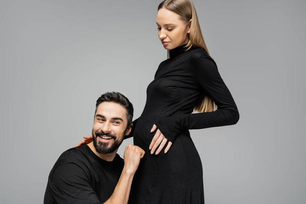 Cheerful bearded man in t-shirt knocking on belly of fashionable and pregnant wife in black dress and standing together isolated on grey, growing new life concept, funny, father to be - Photo, Image