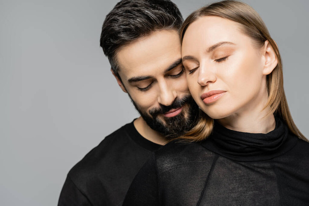 Portrait of relaxed and bearded man in black t-shirt standing next to wife with fair haired wife with natural makeup and closed eyes isolated on grey, husband and wife relationship concept - Valokuva, kuva