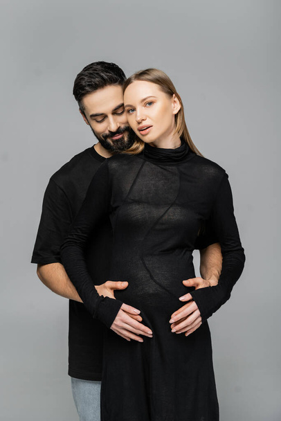 Bearded man in black t-shirt touching belly of stylish and pregnant fair haired wife in dress and standing together isolated on grey, concept of pregnancy and birth of child - Photo, Image