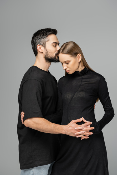 Bearded husband in t-shirt kissing and touching belly of stylish pregnant wife in black dress and standing isolated on grey, concept of expecting parents, happy family  - Φωτογραφία, εικόνα
