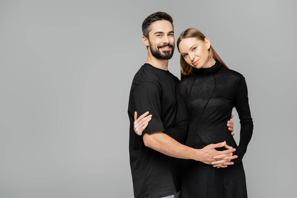 Positive and bearded husband in t-shirt looking at camera while hugging stylish pregnant wife in black dress isolated on grey, new beginnings and parenting concept  - Foto, immagini