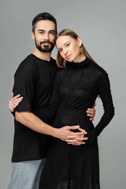 Stilvolle schwangere Frau in schwarzem Kleid, die in die Kamera blickt, während sie die Hand eines bärtigen Ehemannes im T-Shirt hält, isoliert auf grau, Neuanfang und Erziehungskonzept  - Foto, Bild
