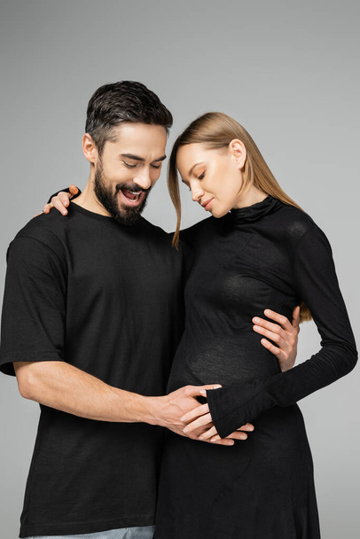 Opgewonden man met baard in t-shirt raakt buik en kijkt naar trendy zwangere vrouw in jurk terwijl ze samen staan geïsoleerd op grijs, nieuw begin en ouderschap concept  - Foto, afbeelding
