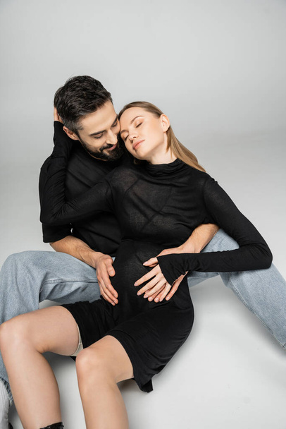 Smiling and bearded man in t-shirt and jeans hugging fashionable and pregnant wife while sitting together on grey background, new beginnings and parenting concept  - Photo, Image