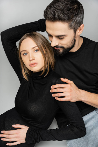 Portrait of bearded husband in t-shirt and jeans hugging stylish and pregnant woman in black dress while sitting on grey background, new beginnings and parenting concept, husband and wife   - Valokuva, kuva
