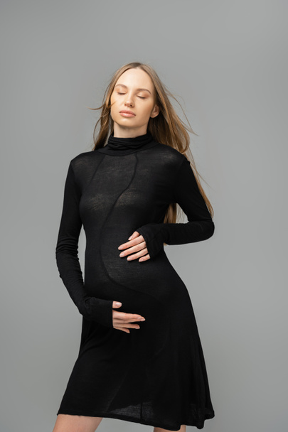 Relaxed fair haired and pregnant woman in stylish black dress closing eyes and touching belly while standing isolated on grey, new beginnings and maternity concept, mother-to-be - Zdjęcie, obraz