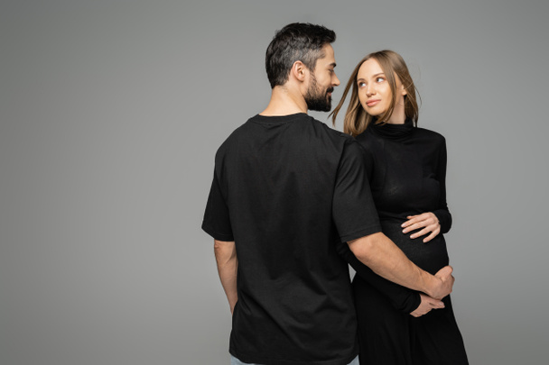 Smiling and bearded man in black t-shirt touching belly of pregnant stylish wife and looking at each other while standing isolated on grey, new beginnings and anticipation concept   - Foto, imagen