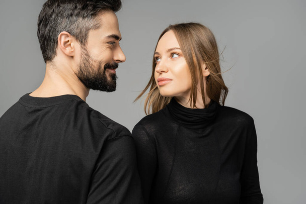 Portrait of stylish fair haired woman looking at smiling and bearded husband in t-shirt while standing together isolated on grey, new beginnings and anticipation concept   - Photo, Image