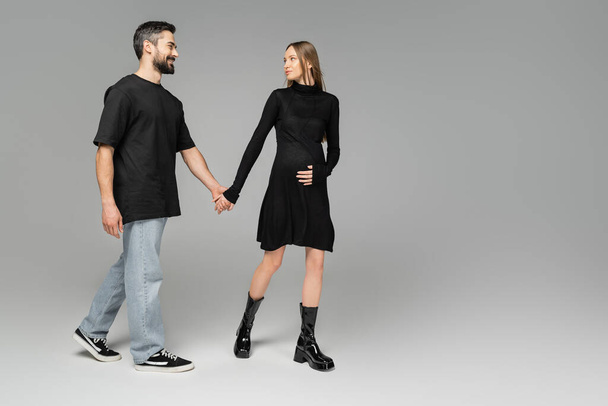 Full length of fashionable pregnant woman in dress holding hand of cheerful husband in jeans and black t-shirt while walking on grey background, new beginnings and anticipation concept   - Photo, Image