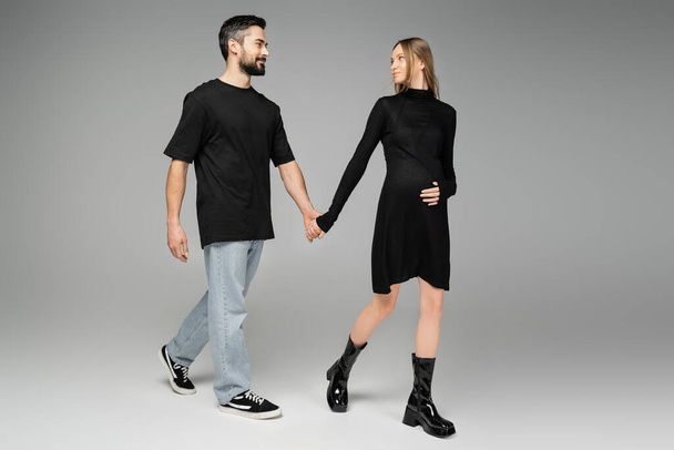Full length of smiling man in jeans and black t-shirt holding hand of stylish pregnant wife in dress and walking on grey background, new beginnings and anticipation concept, expecting parents  - Photo, Image
